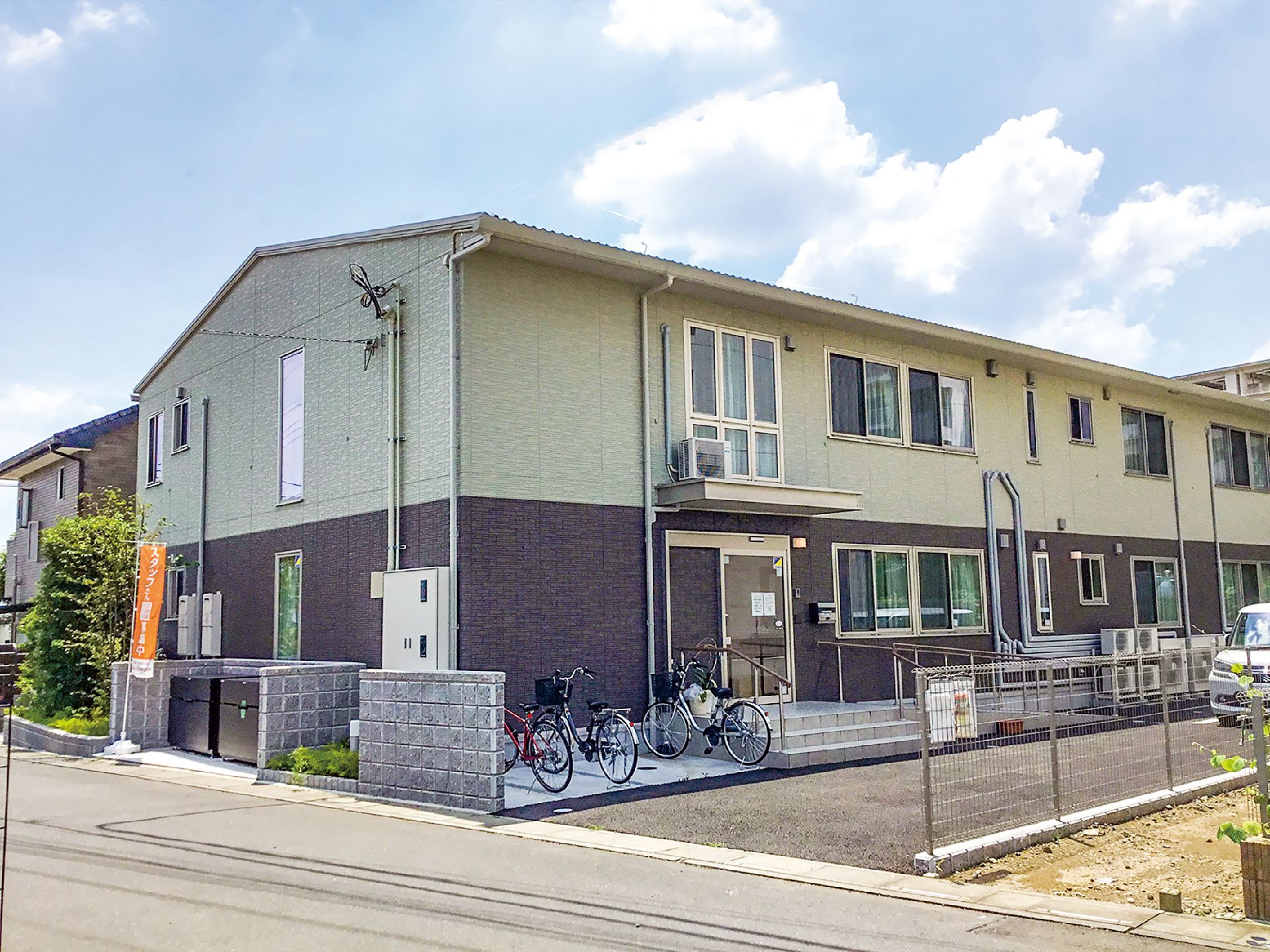 玄関には手すり付きのスロープと階段が付いた2階建ての建物。グレーとブラウンで落ち着いた雰囲気。駐車場もある。
