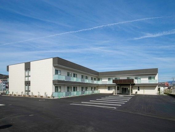 白線が弾かれた広い駐車場が付いた建物は白でエル字型。角フロアにベランダが付いている。