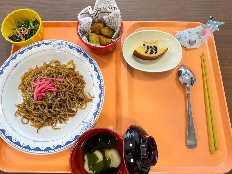 紅ショウガが添えられた焼きそばやこいのぼりが書かれたバームクーヘン、青菜の和え物、汁物などが並べられている。