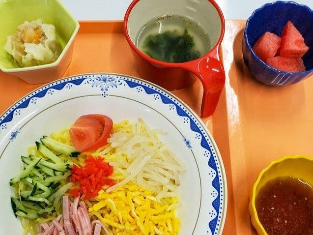 カットスイカやわかめスープ、冷やし中華、野菜の和え物などが並べられたオレンジのトレー。