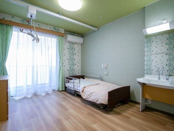 エアコンや介護ベッドが置かれたフローリングの部屋には介護リフトがあり、レースカーテンとカーテンが付いた窓がある。