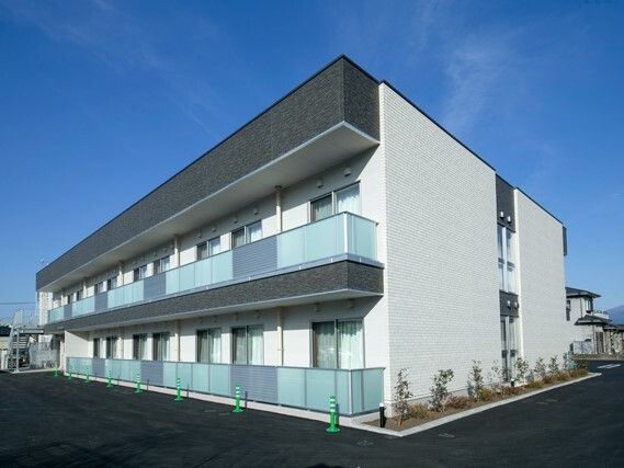角フロアにベランダが付いた白を基調とした２階建ての建物は日当たり良好。敷地内には草木が植えられている。