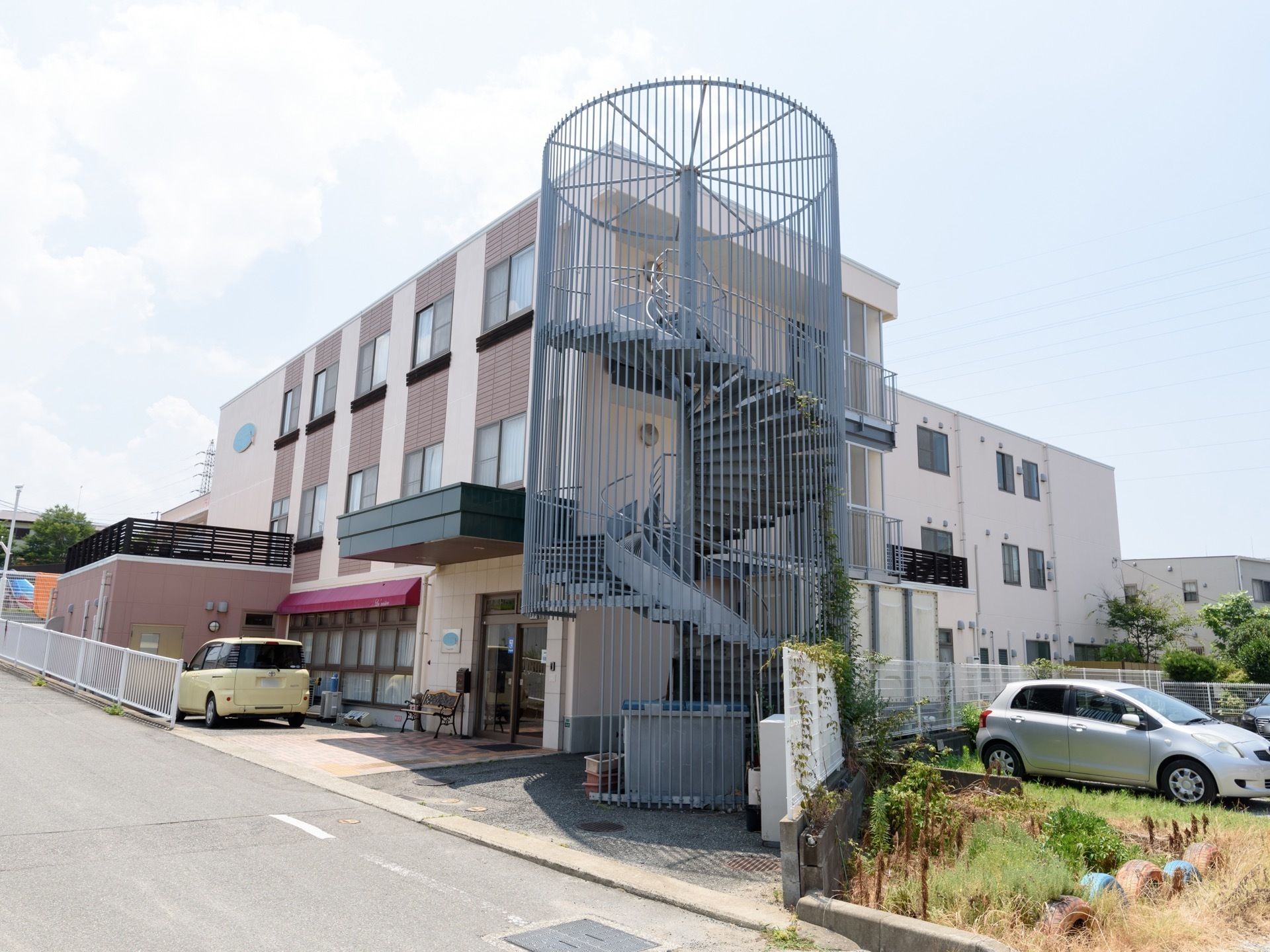 エントランスの横に、らせん階段が外壁に沿って設置されている３階建て施設の外観