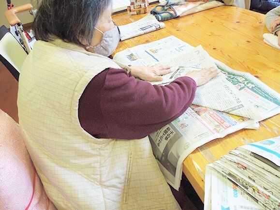 共有スペースの木製のテーブルの上で新聞紙を折っている女性入居者。女性入居者はベストを着て、マスクをつけている。