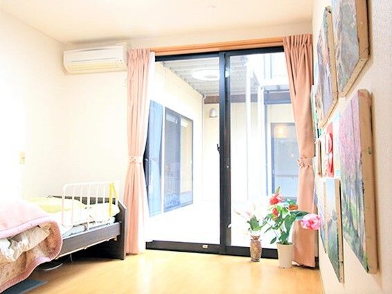 寝具付きのベッドが壁際に置かれ、大きな窓にはカーテンが付き、壁にはたくさんの絵画が飾られたフローリングの居室。