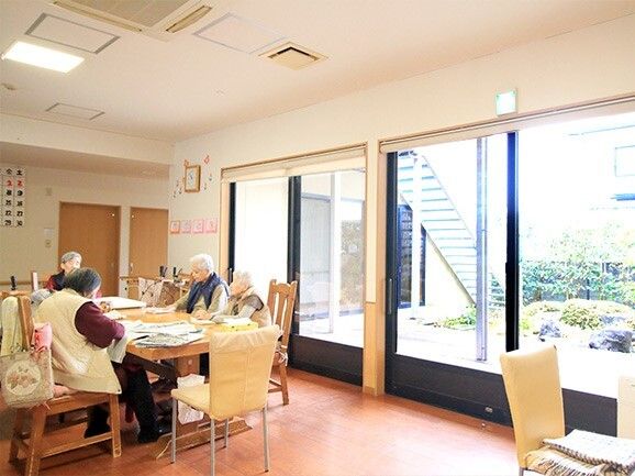 大きな窓が付いた明るいリビングダイニング。木製のダイニングテーブルには4名の女性入居者がいて、思い思いのことをしている。