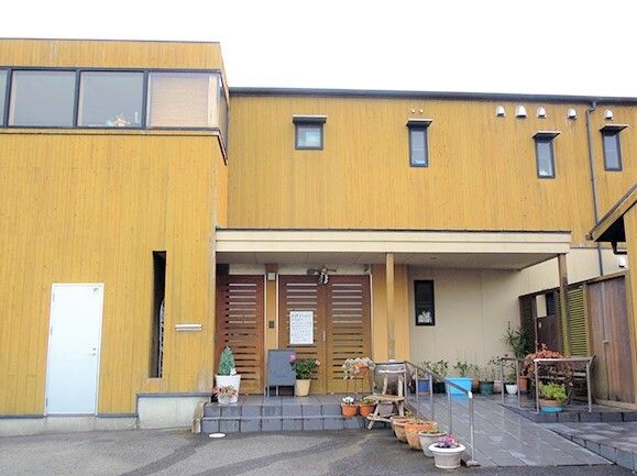 木目調の外壁が明るい雰囲気の2階建ての建物。手すり付きのスロープが付いた玄関にはたくさんのプランターも並べられている。