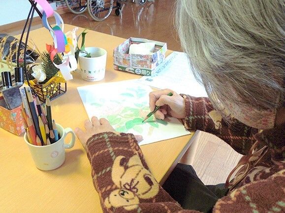 白髪の高齢女性が熱心に塗り絵をしている様子。テーブルの上には色鉛筆がたくさん入ったコップやチラシで作ったゴミ箱が置かれている。