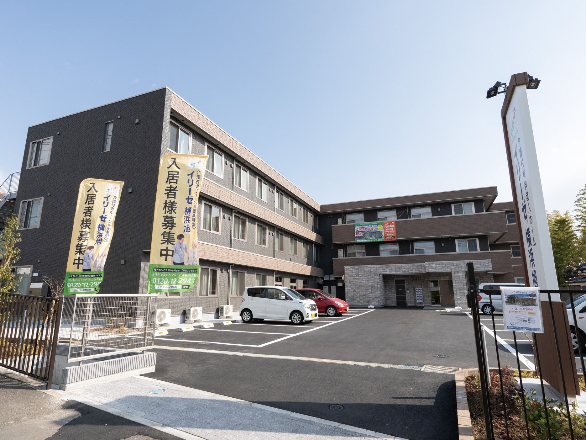ダークブラウンの3階建ての建物はエル字型で大きな駐車場があり、敷地の周りにフェンスが立っている。