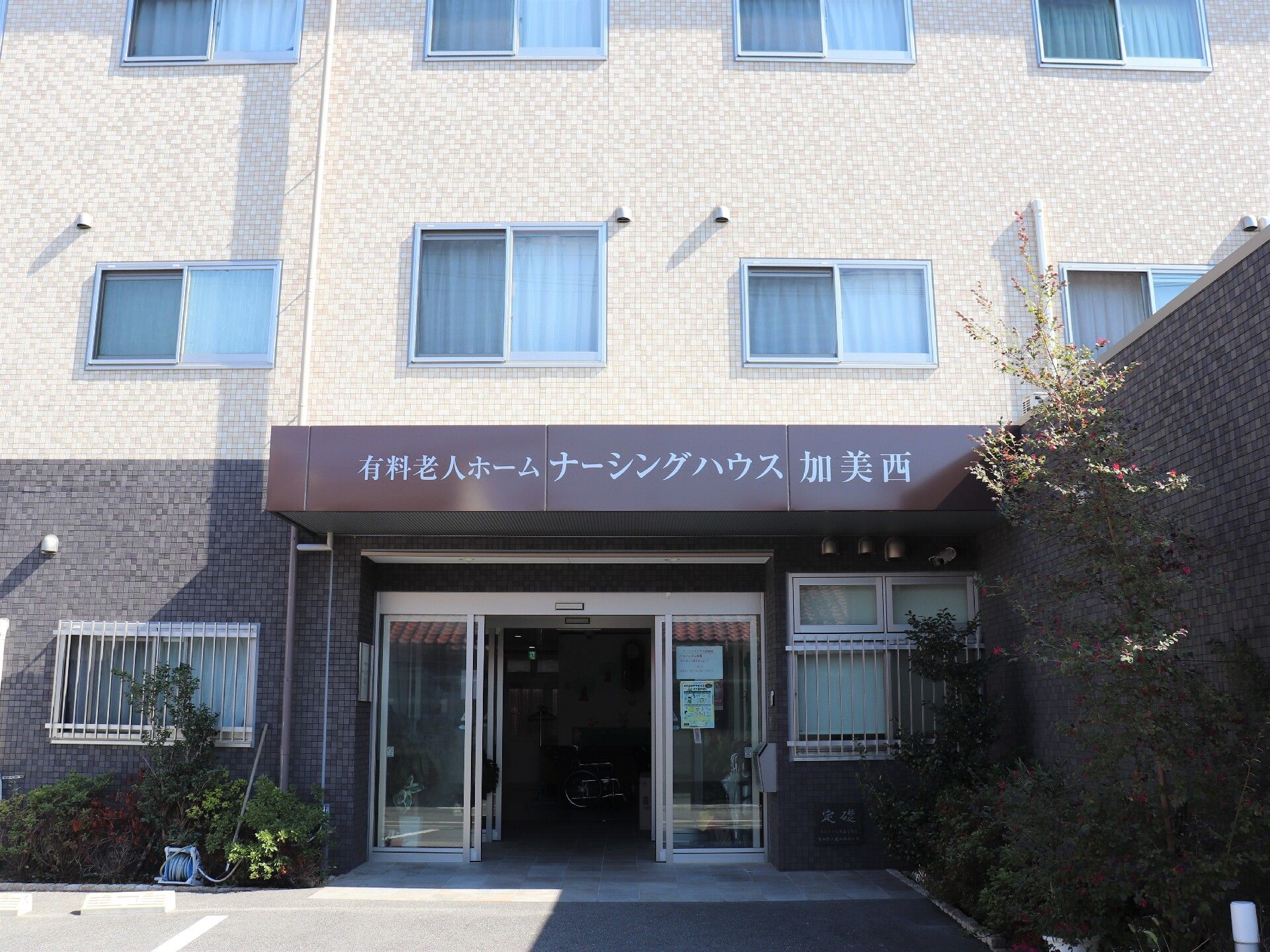 ３階建ての建物はベージュとブラウン系で駐車場もある。玄関部分には屋根が付き、建物の周辺には緑も植えられている。