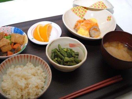 白いご飯に味噌汁、いんげんの和え物に野菜の煮物、オレンジ、卵料理などが並んだトレー。