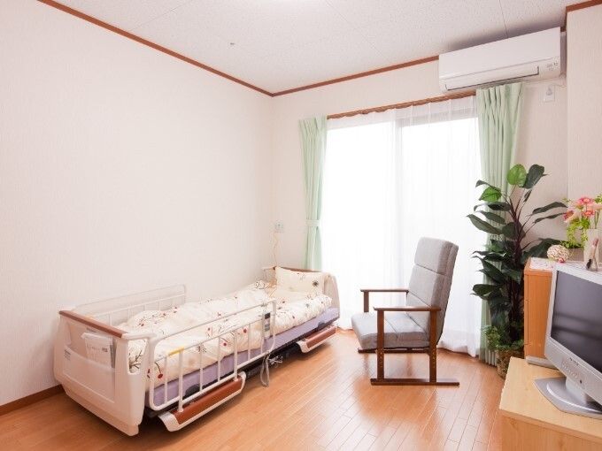 エアコンが付いたフローリングの部屋。大きな窓があり、寝具付きのベッドが置かれ、テレビやイスもある。