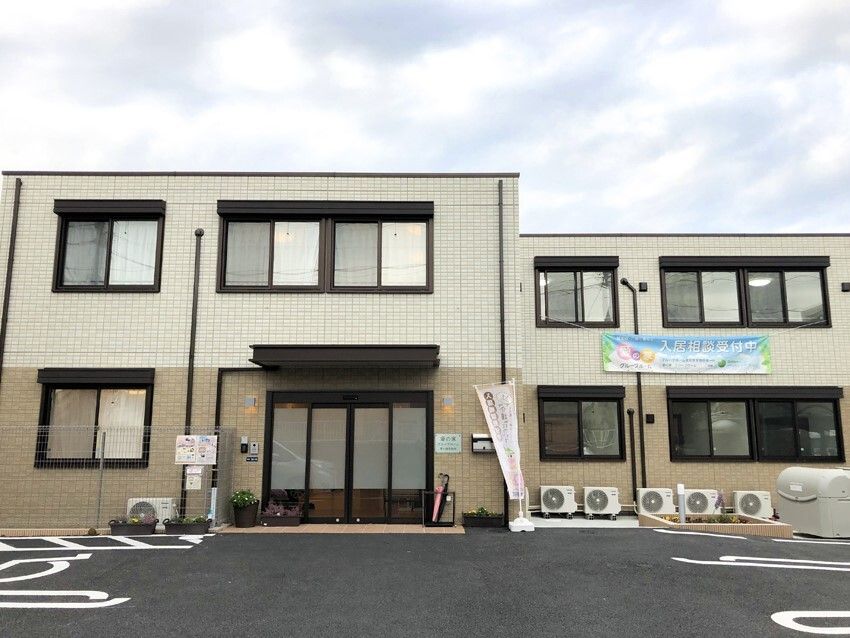 白とベージュの2階建ての建物の前には駐車場もあり、車いすマークが描かれているスペースもある。