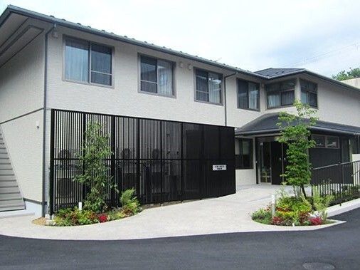 2階建ての建物は横に長く、屋根が付き、建物周辺には黒い柵のようなものがあり、玄関には車寄せが付き、緑も植えられている。