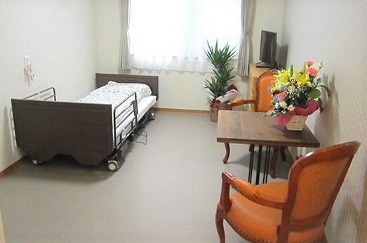 寝具付きのベッドが窓際に置かれ、チェストの上にはテレビがあり、テーブルの上にはフラワーアレンジメントも飾られている。