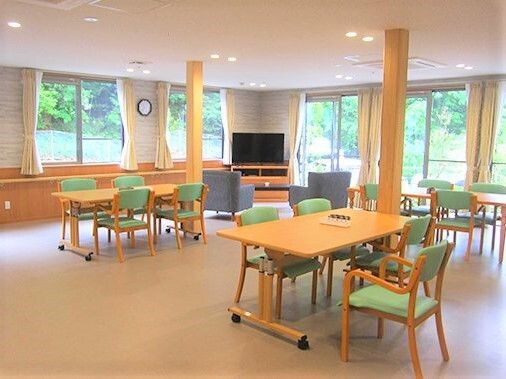グリーンの椅子がセットされたダイニングテーブルが複数並んだ日当たりのいい食堂。テレビやソファが置かれたリビングスペースもある。