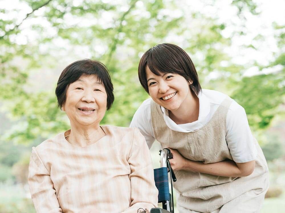 ベージュのエプロンを付けた女性スタッフに付き添われて屋外で笑顔で過ごしている車いすの高齢。