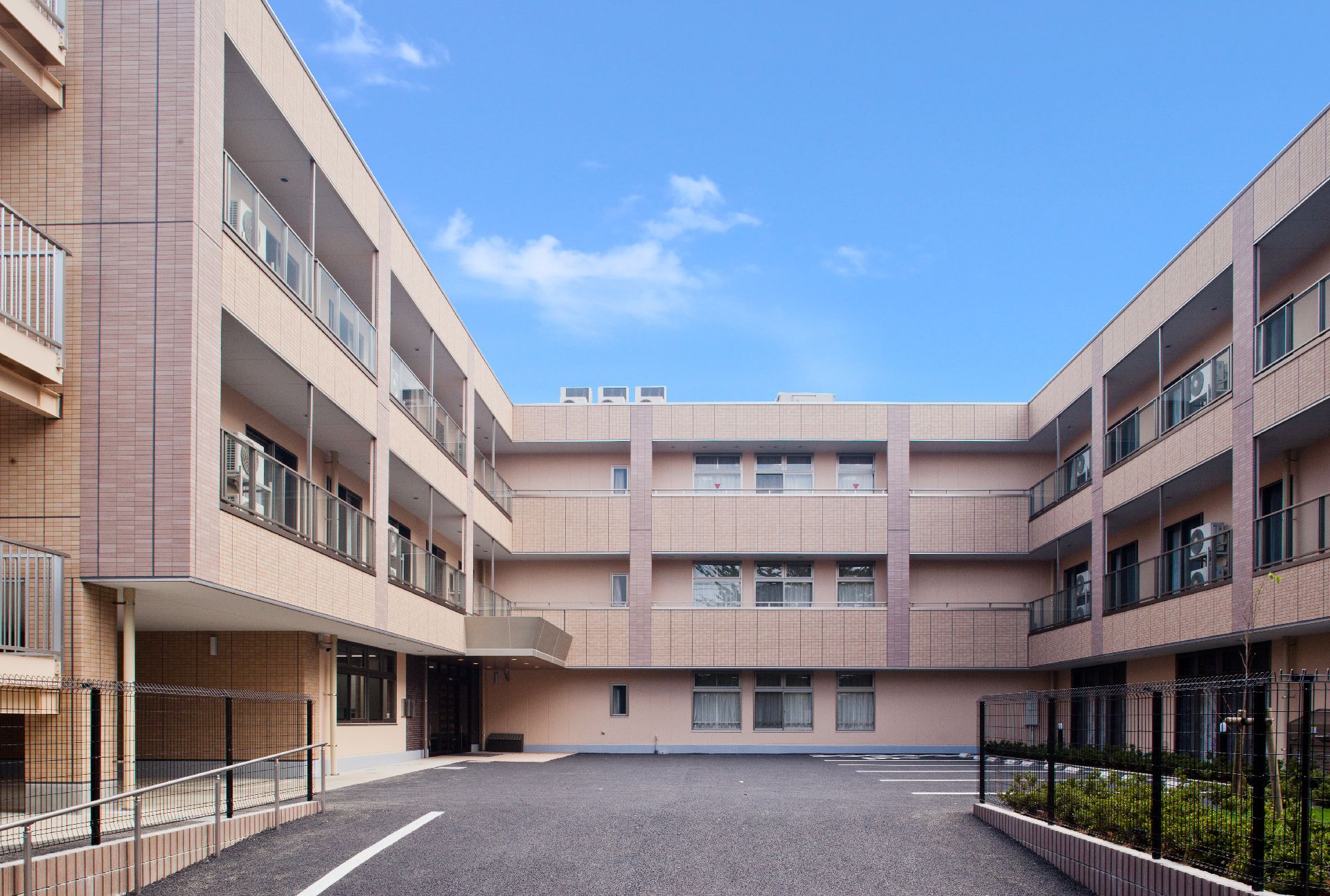 緑に囲まれ３階建ての建物。各フロアにベランダがあり、道路に面したベージュの建物。