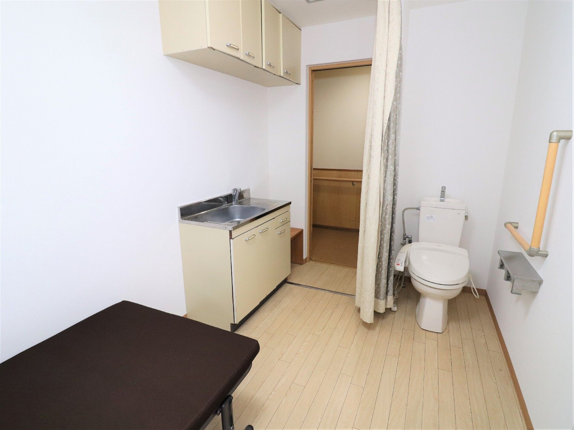 トイレやベッド、流しが付いたフローリングの部屋。トイレはカーテンで開け閉めするタイプで手すりもある。