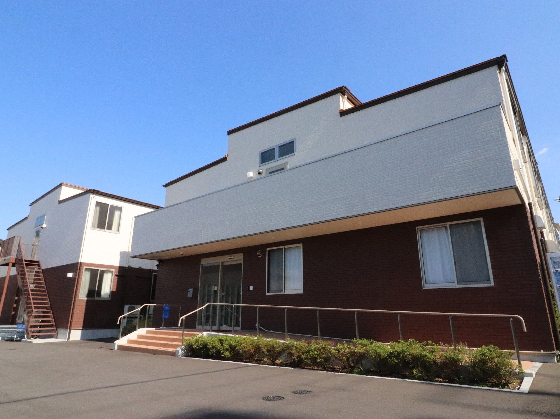 玄関周りには手すり付きのスロープが付き、日当たり良好。2階建ての建物は白とブラウンで落ち着いた雰囲気。