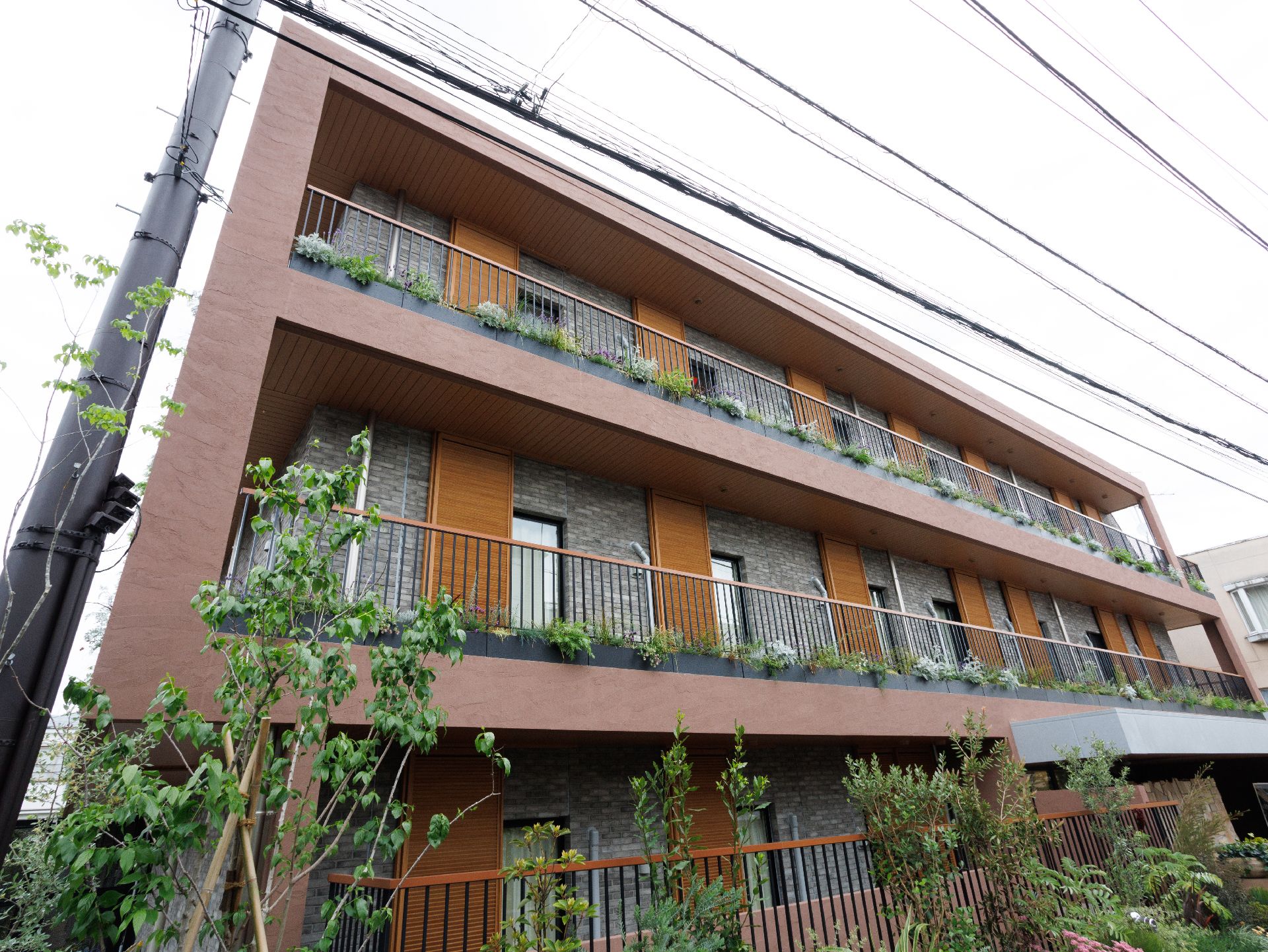 ブラウン系の3階建ての建物の各フロアにベランダがあり、ベランダや建物の草木が植えられている。