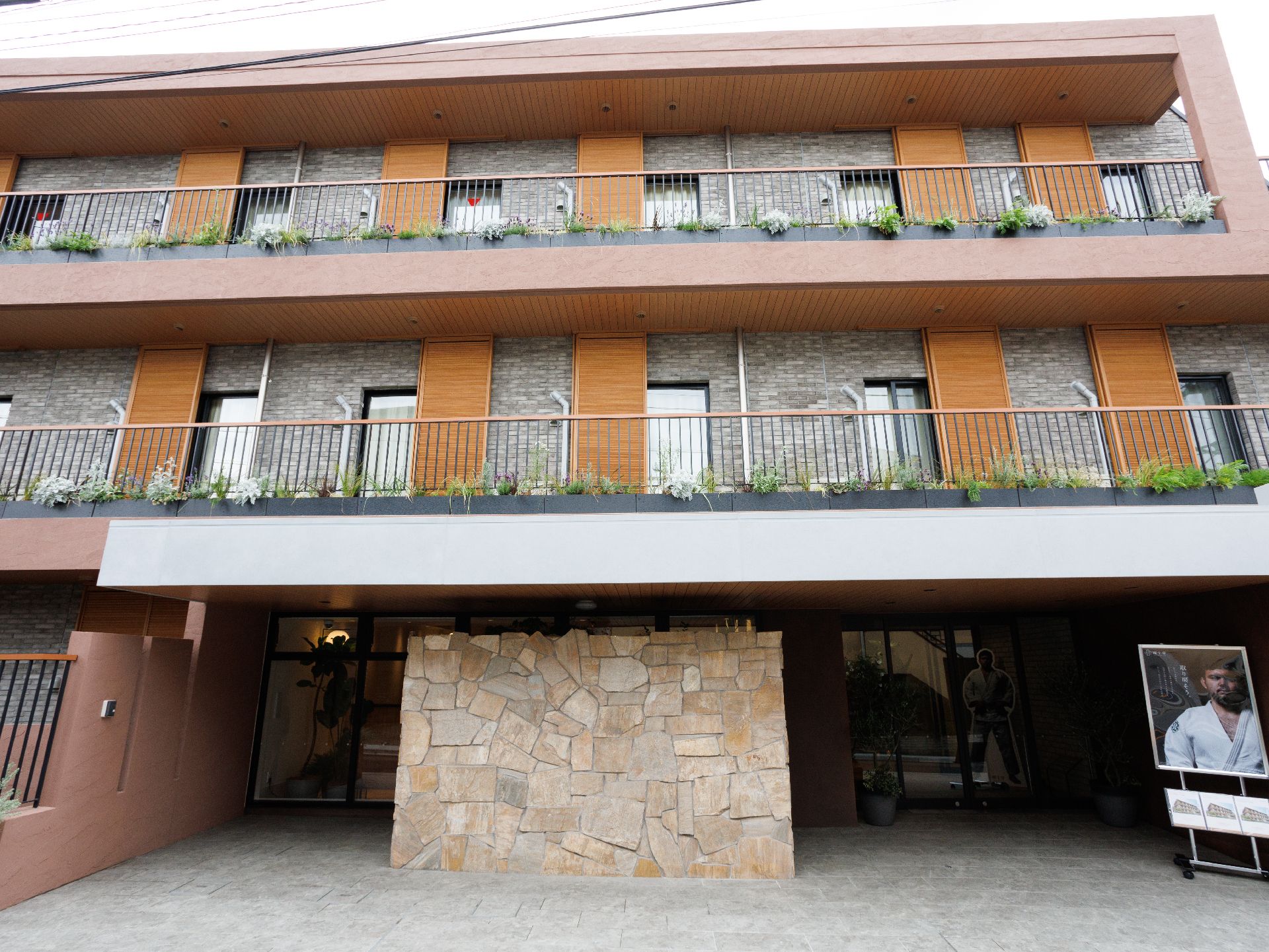 各フロアにベランダがついたブラウン系の３階建ての建物の周りには草木が植えられている。