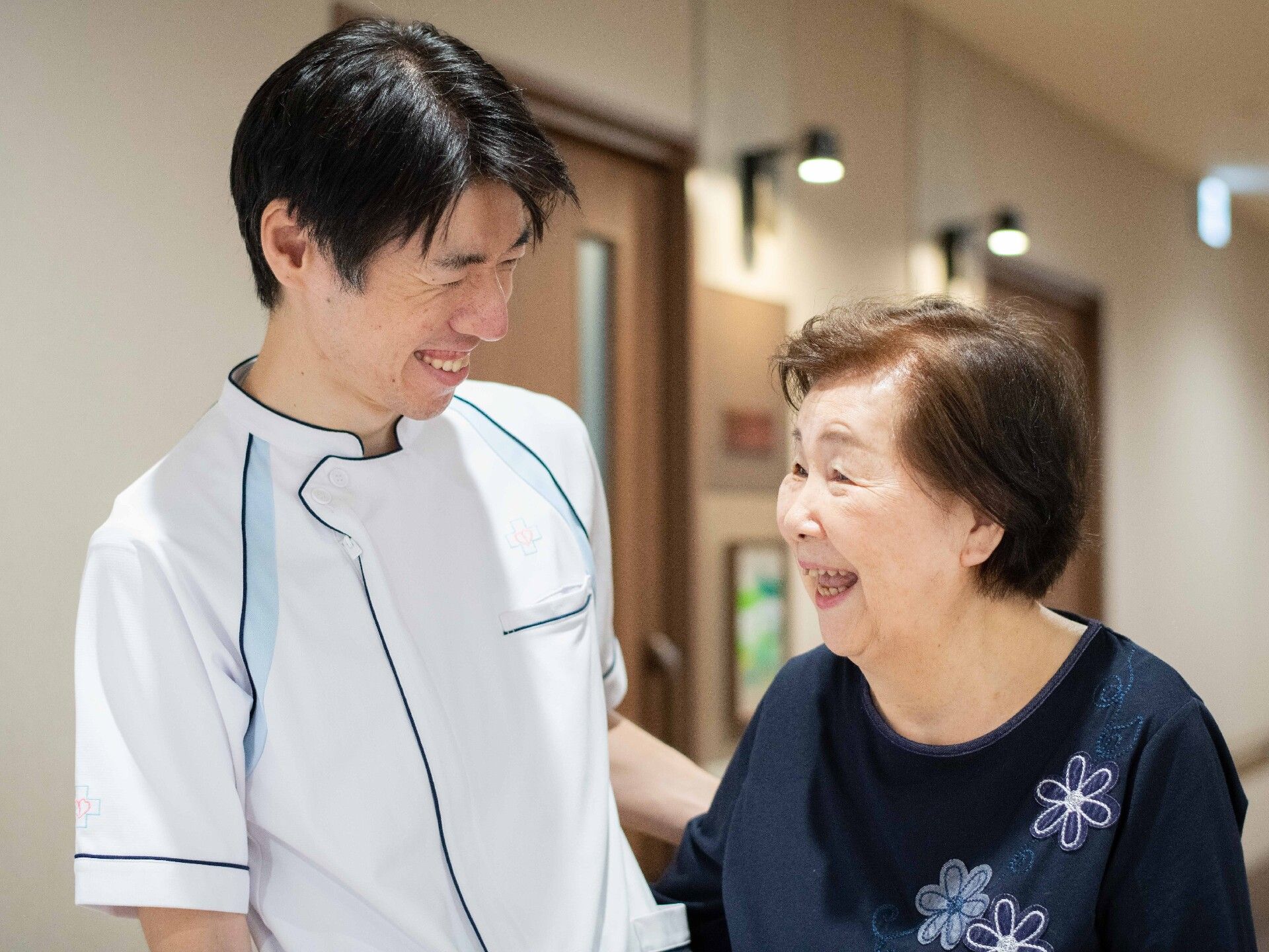 白衣を着た女性スタッフが車いすの高齢女性の車いすを押し、館内を移動している様子。