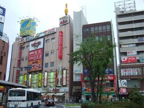 駅前周辺の外観である。駅のロータリーの周りにはビルが並んで建っており、飲食店、ゲームセンターなどの店舗の看板が見られる。
