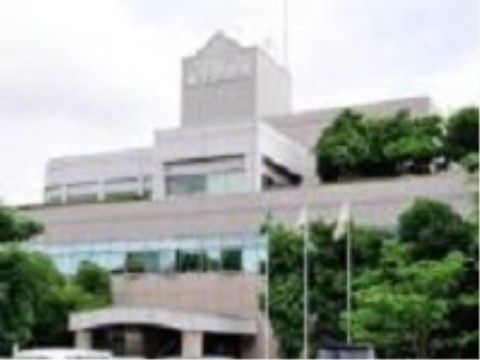 市役所の建物の外観である。建物の壁の色は明るいグレー系で、玄関には大きな屋根が設置され、周囲は緑の木々で囲まれている。