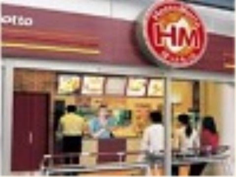 弁当のチェーン店の店舗の正面外観である。大きなガラス戸の内側には客が並んでおり、カウンターでは店員が客に対応している。