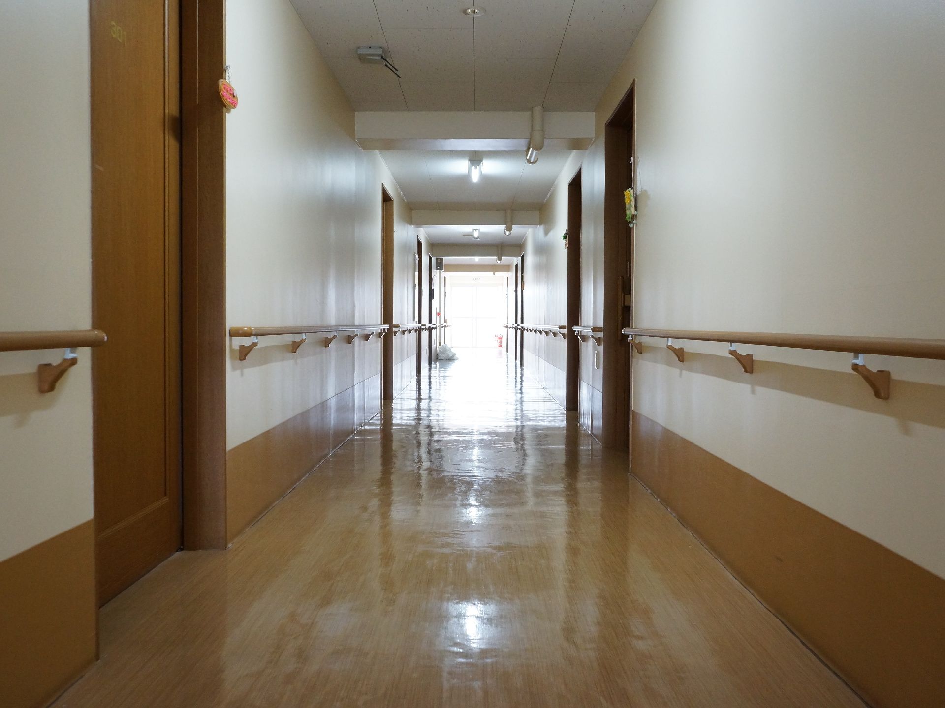 廊下の様子。縦に長く伸びている。奥からは日の光がたくさん入って来ている。居室のドアが並び、壁には手すりが設置されている。