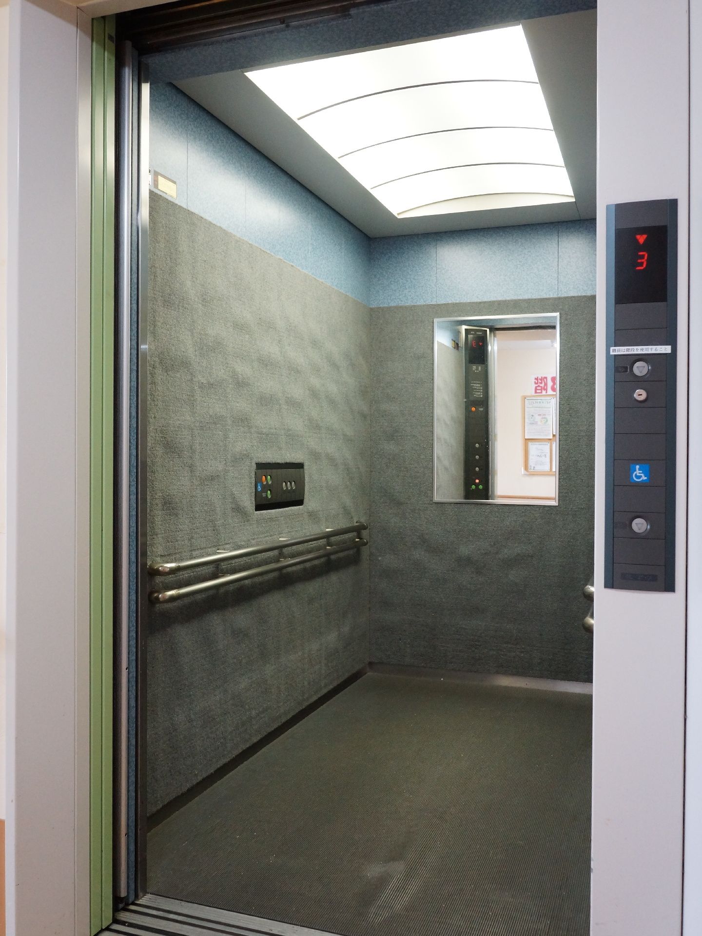 エレベーターを外側から写した様子。ドアが開いて中が見えている。内装はグレーで、奥行きがあり、手すりと鏡が付いている。
