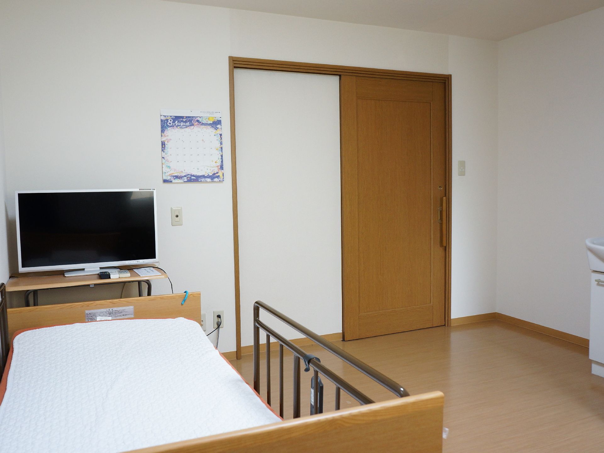 居室を窓側から見た様子。正面に入り口ドアがある。スライド式で木目調。左側にベッドがあり、その前にはテレビが設置されている。