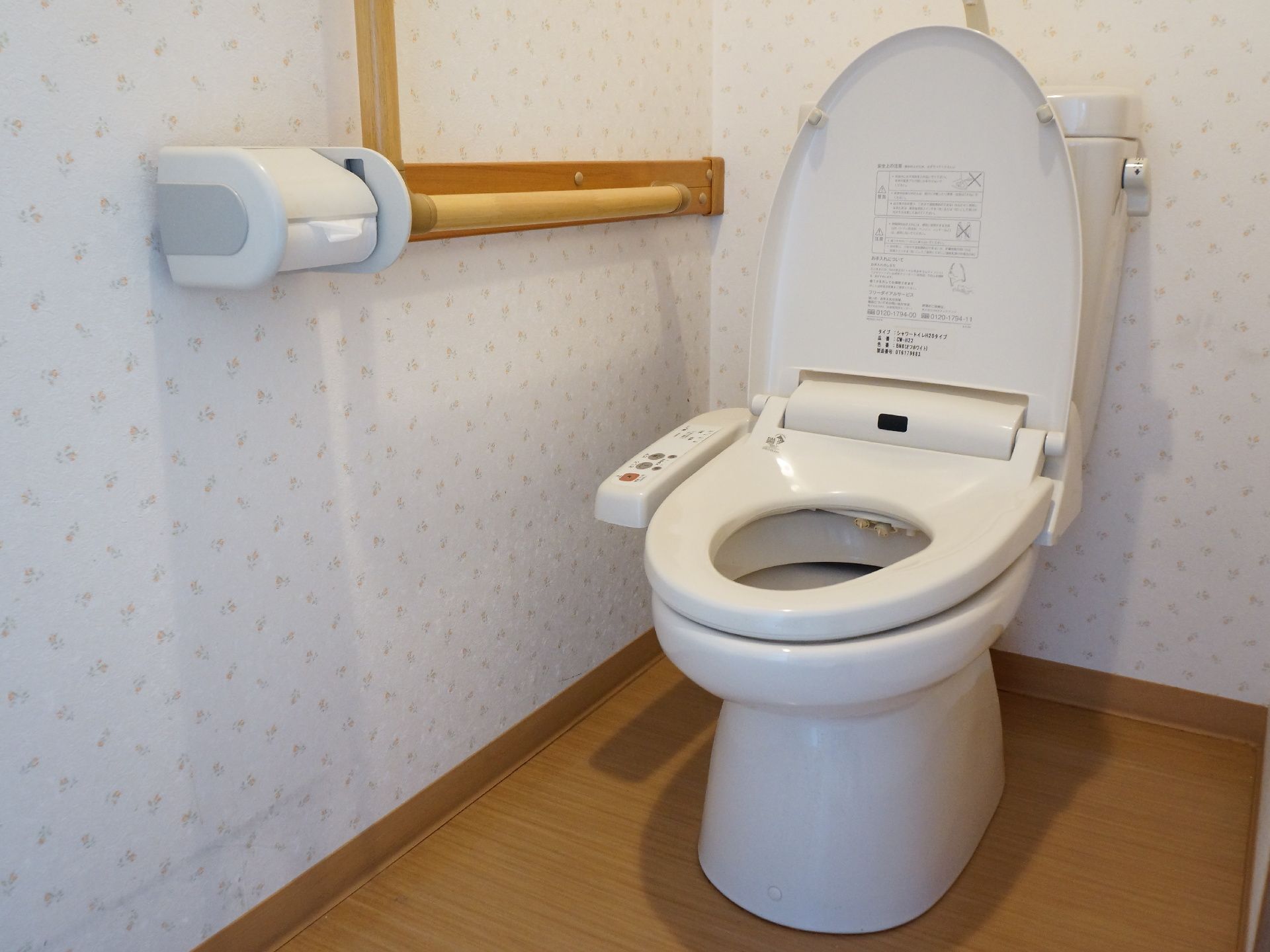 トイレ内部の様子。壁には小花柄のクロスが貼られている。便座は、温水洗浄機能付きの洋式タイプ。手すりが設置されている。