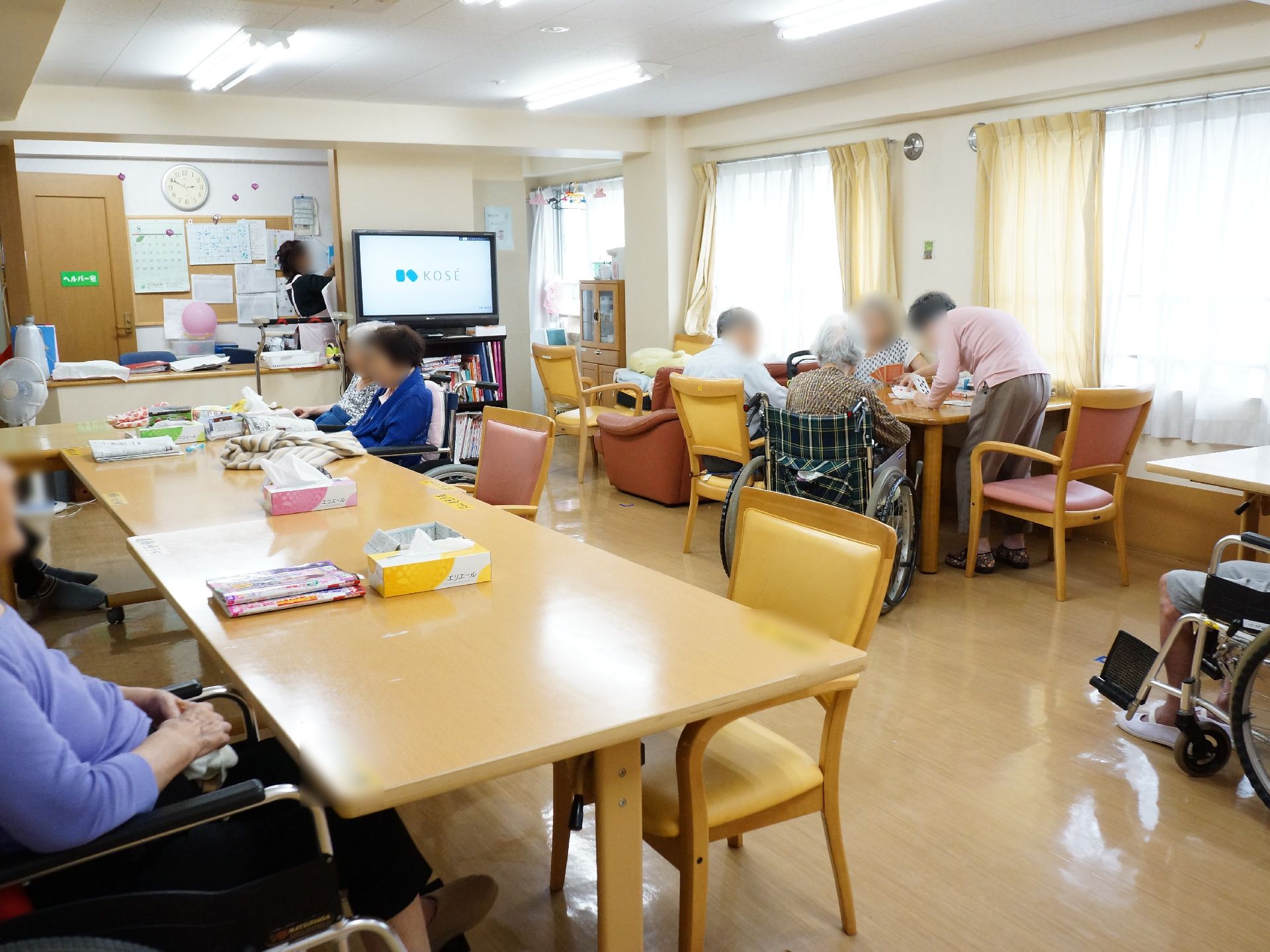複数ある壁際の窓から、明るい日の光が差し込んでいる食堂・談話室。数人の入居者が、テーブルに着いて過ごしている。