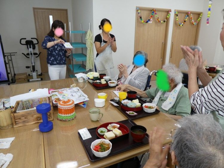 緑の多い屋外で笑顔で誰かに手を振っている車いすの高齢女性と車いすを押しているポロシャツを着た男性スタッフ。