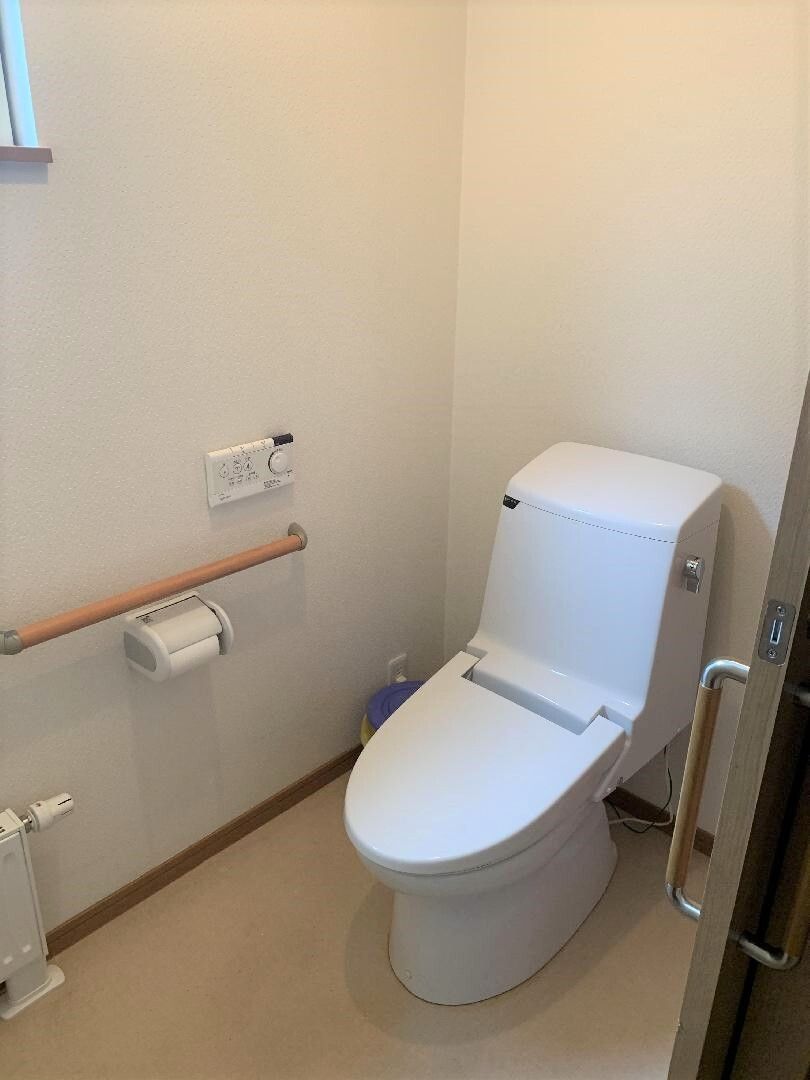 シンプルなデザインの白い便座が付いたトイレ。トイレの右側の壁には操作パネルや手すり。トイレットペーパーホルダーが付いている。