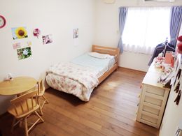 寝具付きのベッドや木製のテーブルセット、チェストが置かれたフローリングの部屋には大きな窓が付き、写真が飾られている。