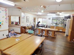大きなダイニングテーブルが置かれたスペース。キッチンには大きな作業台があり、スタッフが作業をしている。テーブルには入居者が一名いる。