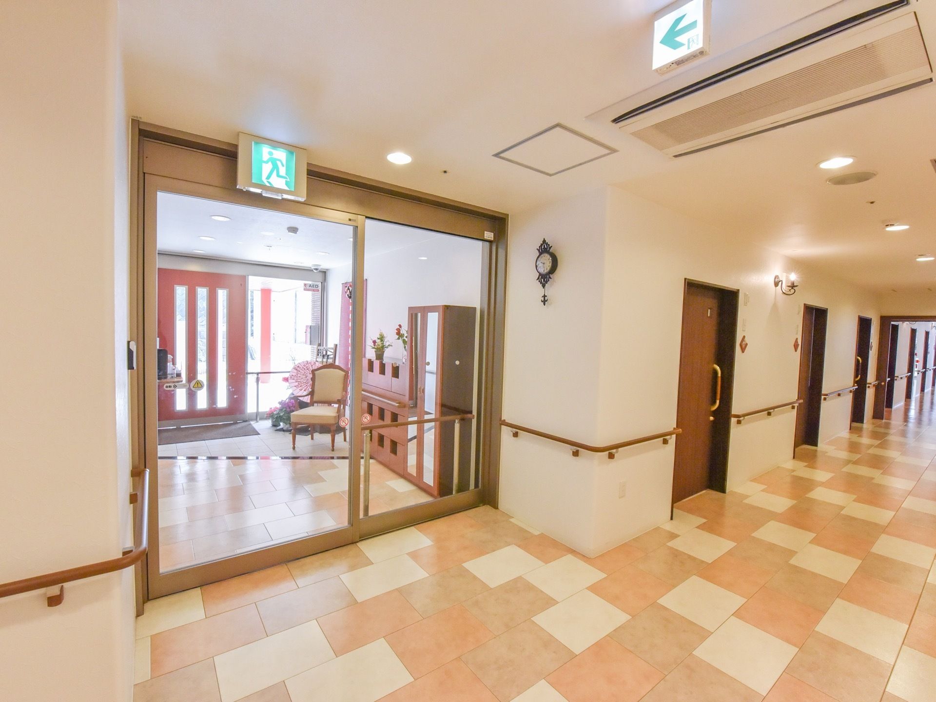 風除室との間には透明の大きな自動ドアがあり、非常口のマークが付いている。風除室には靴箱や椅子が置かれている。