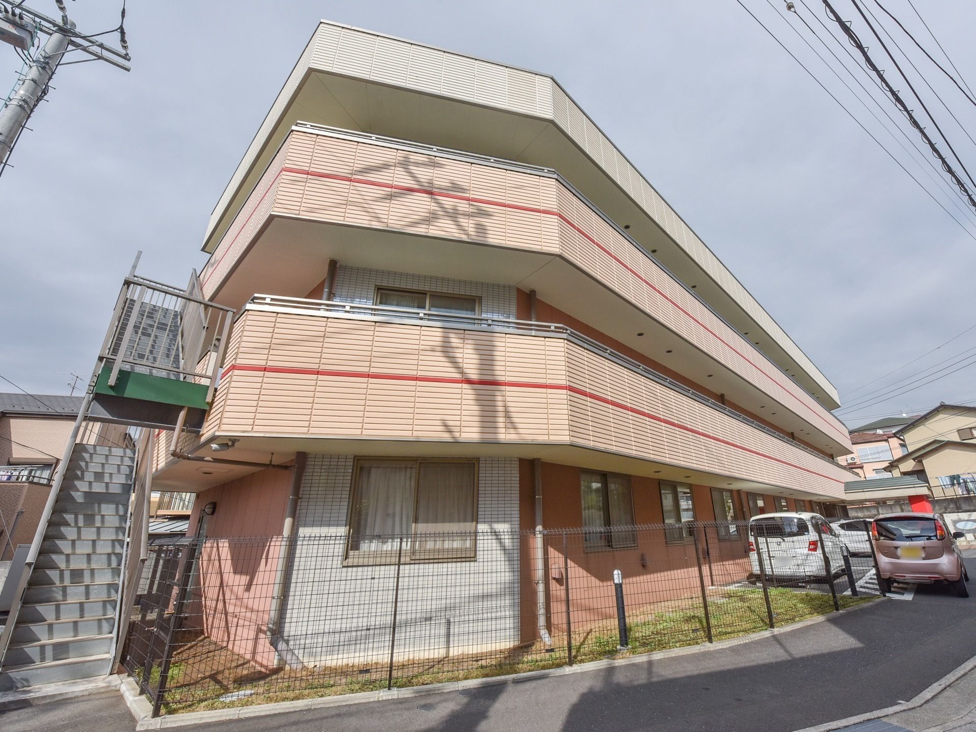 建物の周りをフェンスで囲まれており、その中は芝生が敷かれている。左側に非常階段があり、右側は駐車場である。