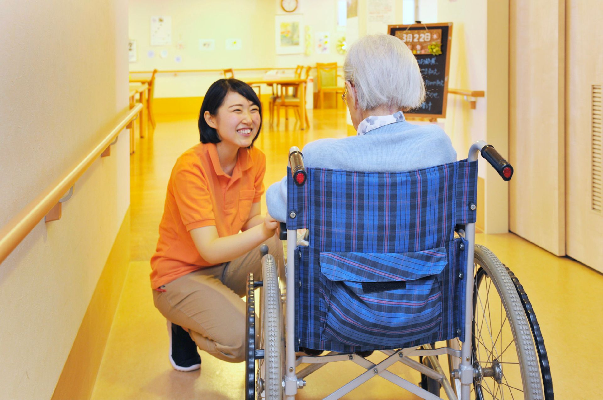 手すり付きの幅の広い廊下で、青いチェック柄の車いすに座った高齢女性にオレンジのポロシャツを着た女性スタッフが笑顔で話かけている。