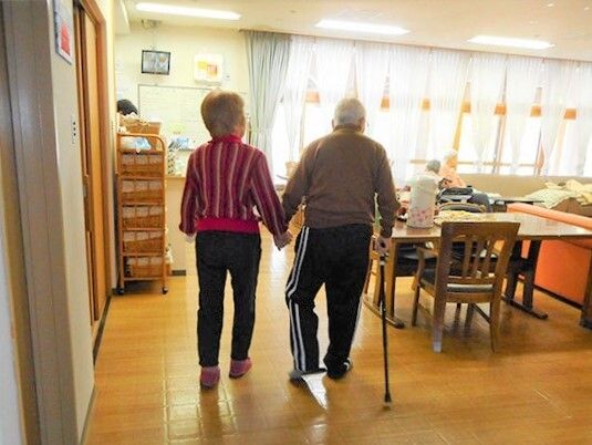 男女の入居者が手をつないで共有スペースを歩いている様子。男性は杖を突いている。