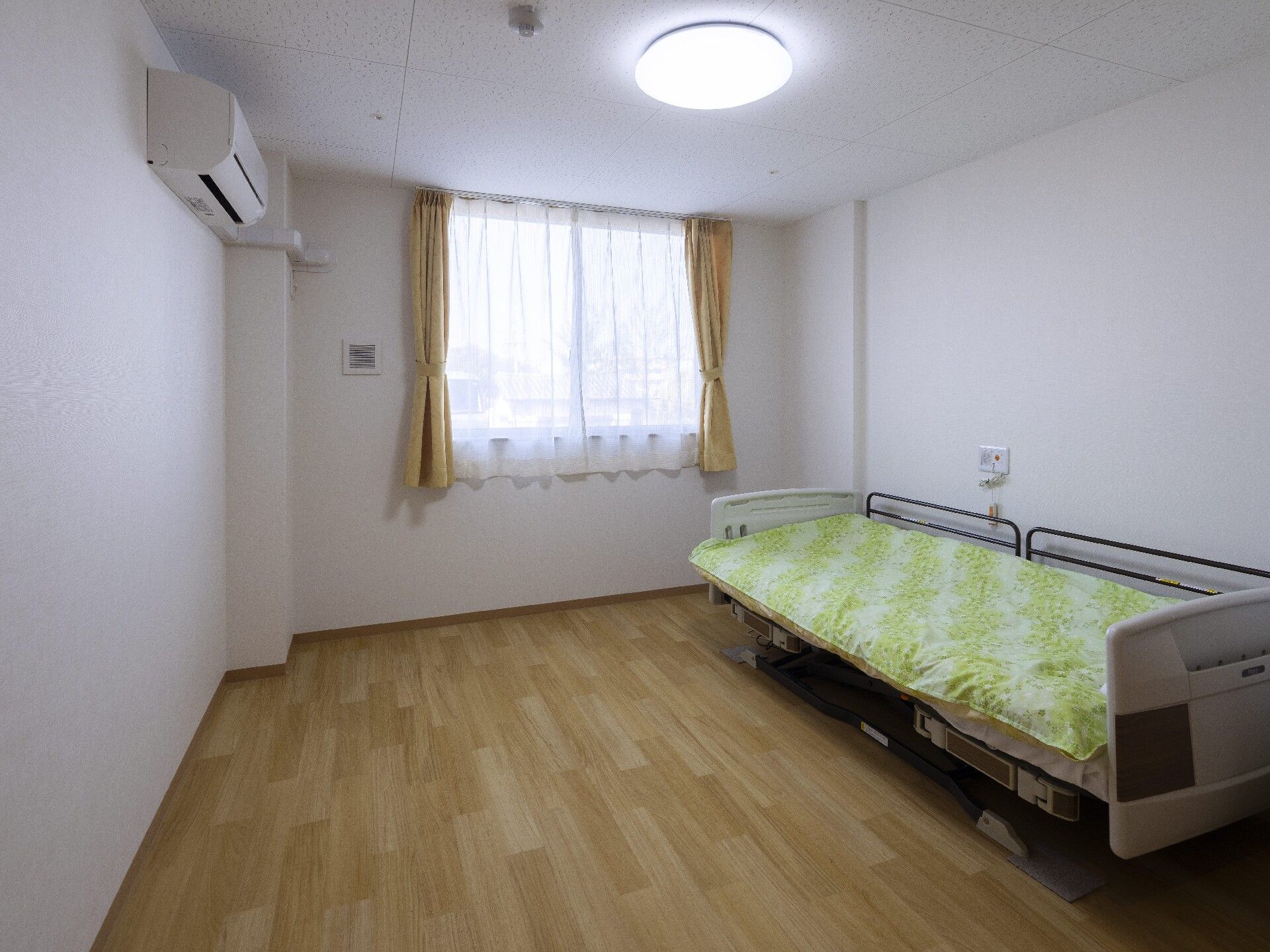 寝具付きのベッドやエアコンが設置されたフローリングの部屋。ベッドサイドには緊急通報装置が付いている。