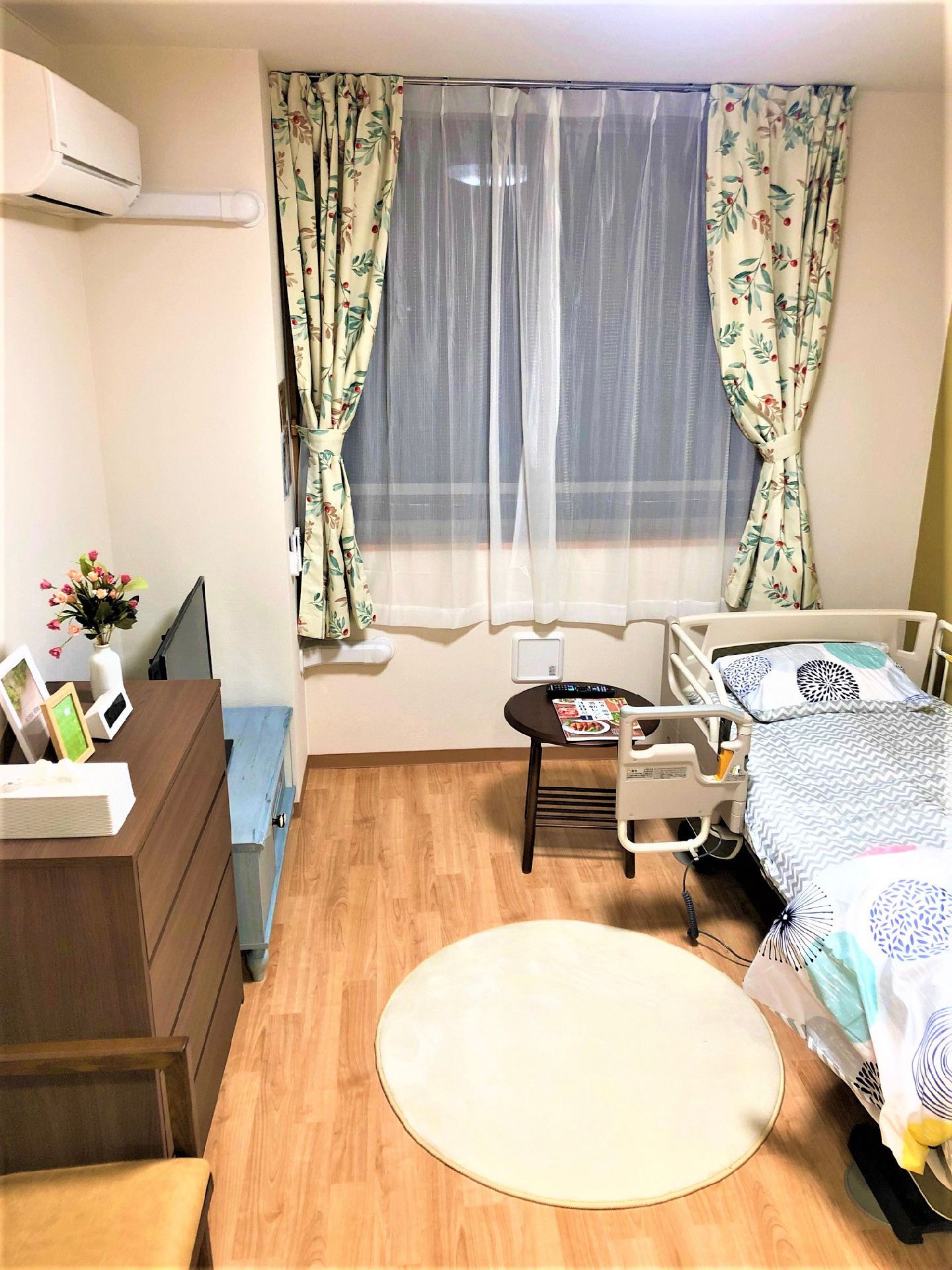 カーテンやレースカーテンが付いた大きな窓があるフローリングの部屋。寝具付きのベッドやテレビ、チェストなどがあり、写真や花も飾られている。