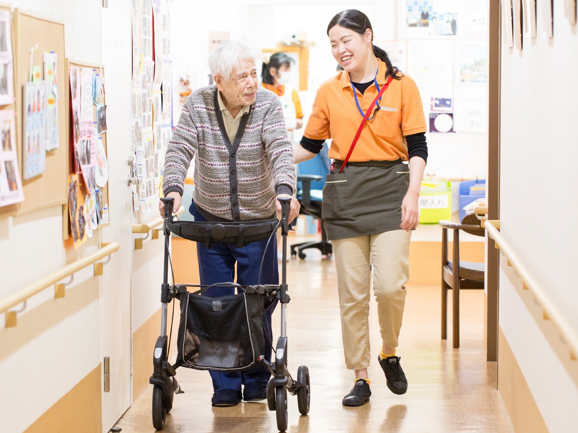 両サイドに手すりが設置された幅の広い廊下を歩く歩行器の高齢男性とそばで笑顔で話かけているオレンジのポロシャツを着た女性スタッフ。