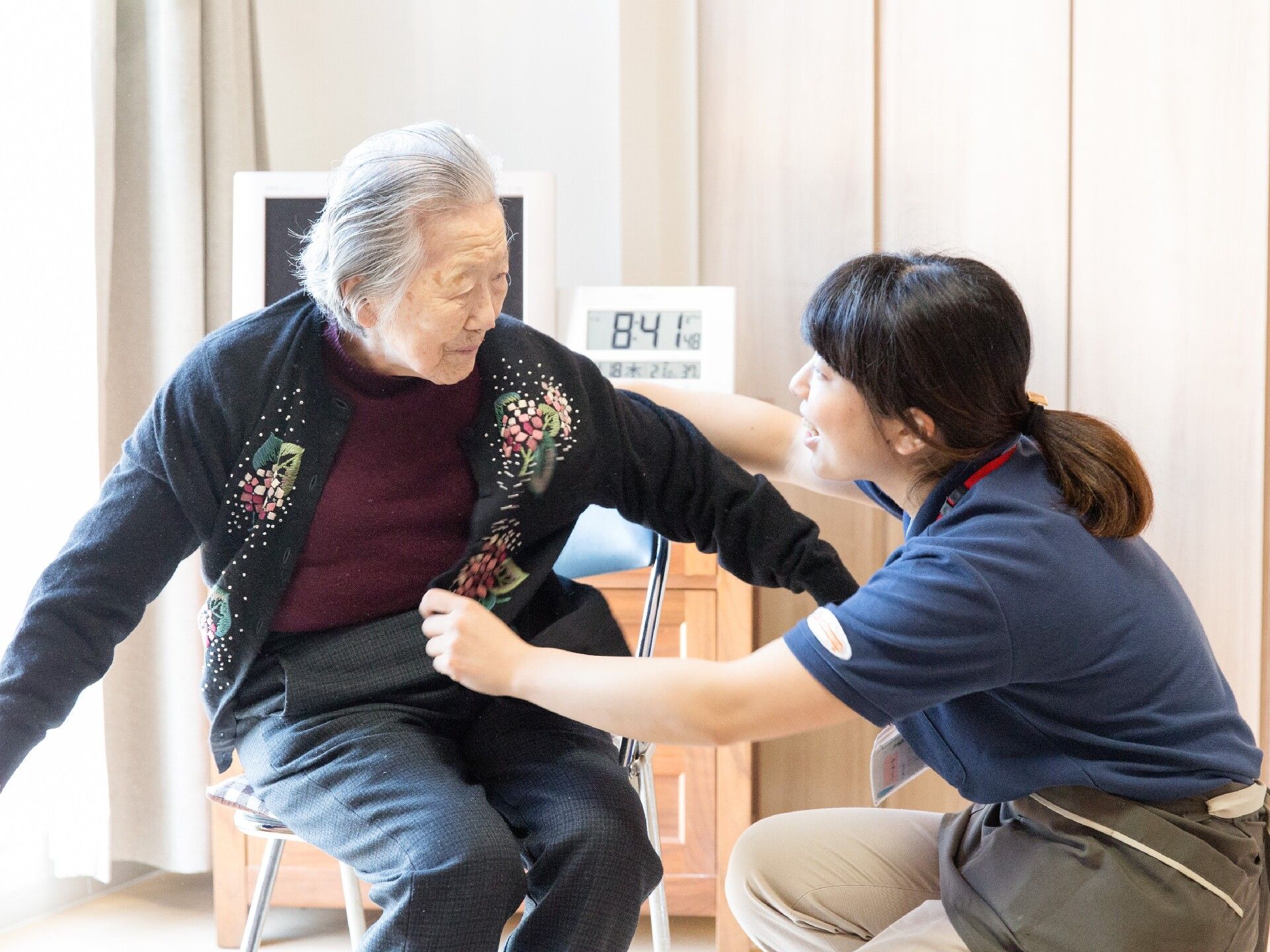 白髪の高齢女性とオレンジのポロシャツを着た女性スタッフが話している。女性は椅子に座り、スタッフは腰を下ろしている。
