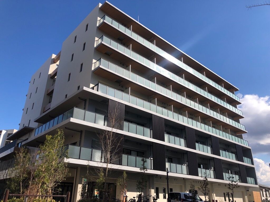 ブラウン系の大きな建物の各フロアにベランダがあり、建物の周りには芝生が敷かれ、玄関のある1階部分は白い外壁。