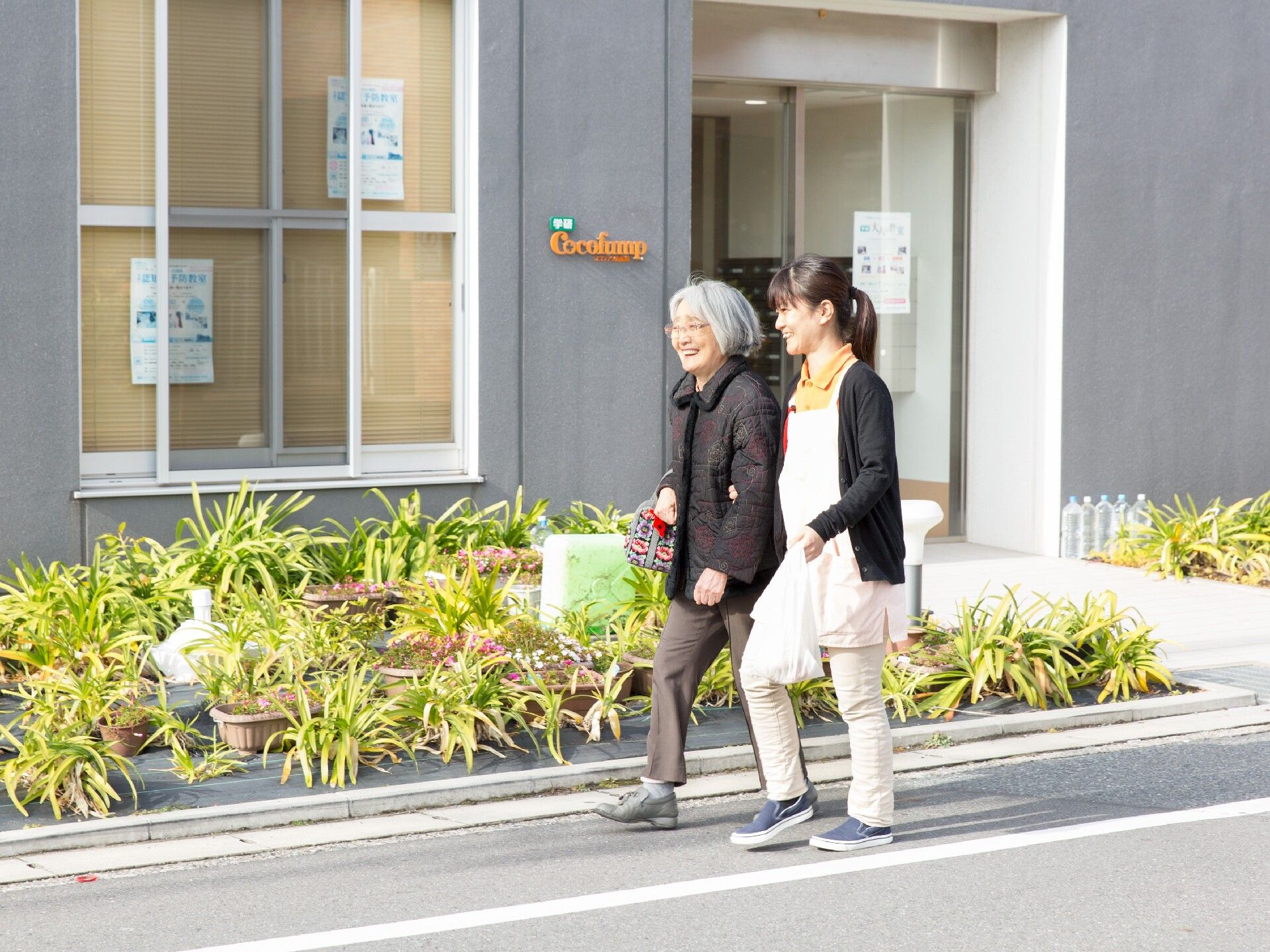 眼鏡をかけた高齢女性と付き添いの若い女性スタッフが外を笑顔で歩いている様子。スタッフはビニール袋を持っている。