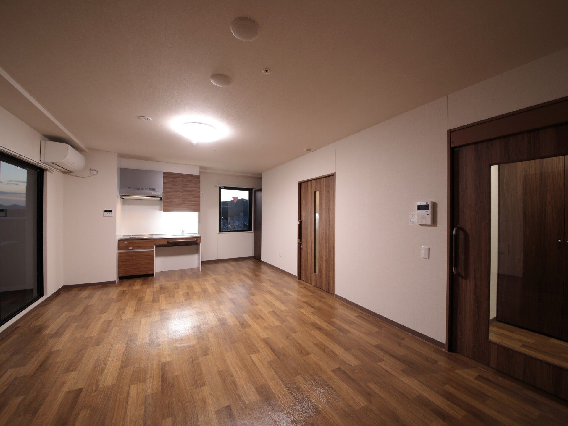 車いす対応のミニキッチンが付いたフローリングの部屋。窓のそばにはラグが敷かれ、一人用の椅子やテーブルが置かれている。